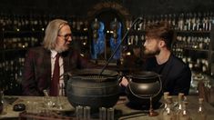 two men sitting at a table in front of an old caulder filled with liquid