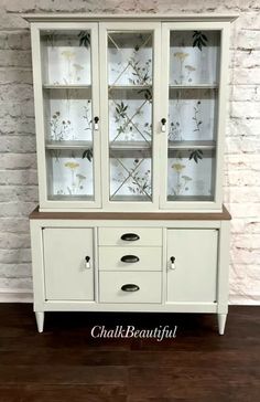 a white china cabinet with glass doors and flowers painted on the front, sitting against a brick wall