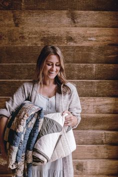 a woman standing in front of a wooden wall holding several blankets and smiling at the camera