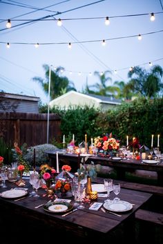 an outdoor dinner table set up with candles, plates and flowers in the center is surrounded by string lights