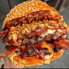 a person holding up a large hamburger with bacon and cheese on the bun, in their hand