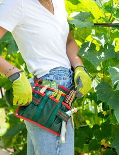 a woman wearing gloves and holding a tool belt in front of some plants with green leaves