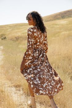 a woman in a brown and white dress walking on a dirt path through tall grass