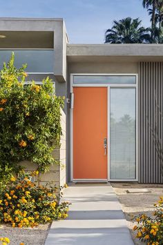 an orange door in front of a house with bushes and flowers on the side walk