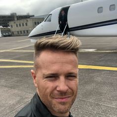 a man standing in front of an airplane on the tarmac with his hair combed back