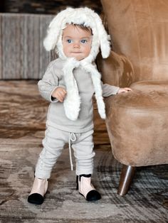 a small child wearing a white hat and grey outfit standing in front of a chair