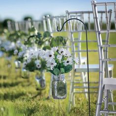 white flowers are in mason jars hanging from the back of chairs at an outdoor ceremony