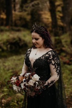 a woman in a black dress holding a bouquet and wearing a tiara standing in the woods