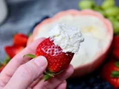 a hand holding a strawberry with whipped cream on it next to grapes and strawberries