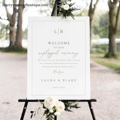 a welcome sign with white flowers and greenery is displayed on an easel in front of trees