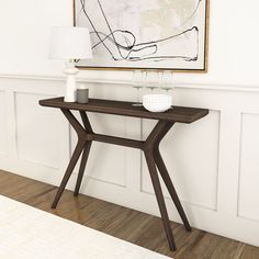 a wooden table with a vase on it in front of a white wall and a painting