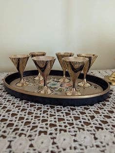 a tray with four glasses on it sitting on a lace tablecloth next to a gold vase