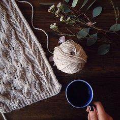 someone holding a cup of coffee next to a knitted blanket and flowers on a table