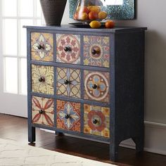 an ornate painted chest of drawers next to a vase with fruit on top in front of a window