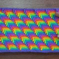 someone is holding up a crocheted rainbow colored dishcloth on a wooden table