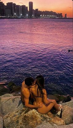 two people are sitting on rocks near the water at sunset, with buildings in the background