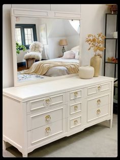 there is a white dresser with a mirror on it and a bed in the background