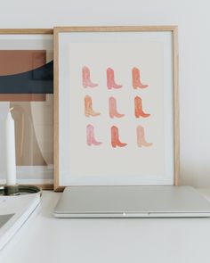 a white laptop computer sitting on top of a desk next to a framed print with pink and orange boots