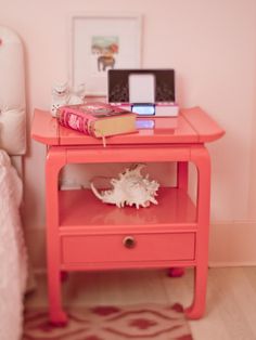 a pink nightstand table with books on it and a white bed in the background,