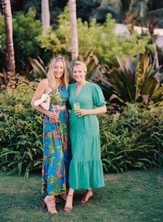 two women standing next to each other in front of some bushes and trees with drinks