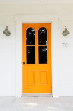 an orange front door on a white house with two light fixtures next to it,