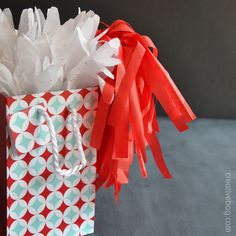 a red and white gift bag with tissue pom - poms on the handles