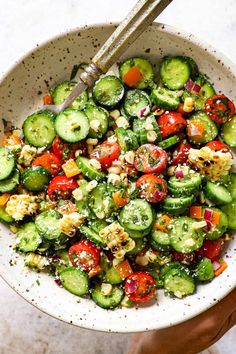 a white bowl filled with cucumbers, tomatoes and other vegetables next to a pair of tongs