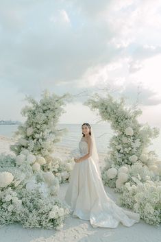 a woman in a wedding dress standing under an arch of white flowers on the beach