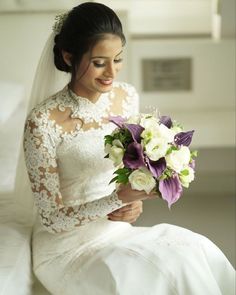 a woman in a wedding dress sitting on a bed holding a bouquet of white and purple flowers