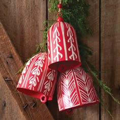 three red and white bells hanging from a wooden wall with pine branches on the side