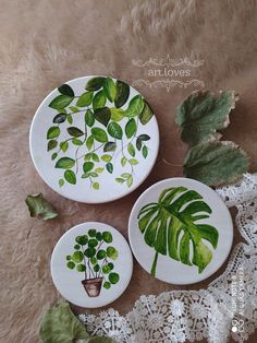 three plates with plants painted on them sitting on a white lace tablecloth next to some leaves