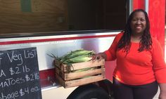 a woman standing in front of a food truck holding a wooden crate filled with vegetables