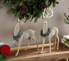 two white deer figurines are standing on a wooden table next to pine cones and evergreen branches