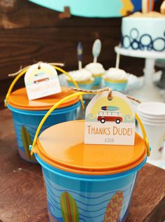 two blue buckets with orange lids are sitting on a wooden table in front of cupcakes