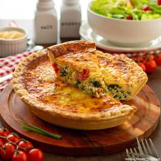 a quiche is sitting on a wooden plate next to a bowl of salad and fork