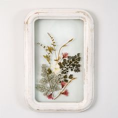 an old white frame with pressed flowers in it on the wall next to a window