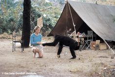 a woman kneeling down next to a black dog on top of a dirt field with a tent in the background