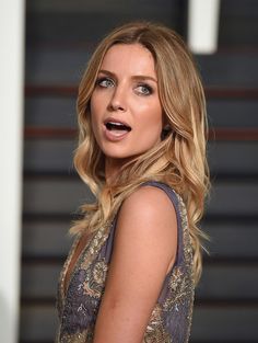 a woman with long blonde hair and blue eyes looks off to the side while standing in front of a dark background