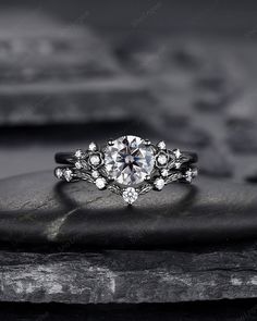 a white diamond engagement ring sitting on top of a black stone slab with rocks in the background