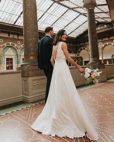 a bride and groom standing together in an indoor area with large columns, tiled flooring and glass ceiling