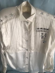 Iconic 80sFashion Designer white silk - Tuxedo/ Western style silkButton down - rhinestone buttons & sleeves wristband Collar has wing tip point Shoulder rhinestone stars - removal shoulder pad 1 sleeve has 3 black line trim w a Isosceles triangle rhinestone patch & round rhinestone w a moon-rhinestone stone Front 1/2 shirt organ pleat -lower 1/2 silk left side of front 1 line of square rhinestone 1 “ “ oval rhinestone 1  upside triangle & square rhinestone 1 large round rhinestone & Embellished Fitted Shirt For Workwear, White Fitted Silk Shirt, Formal Embellished White Tops, Fitted White Silk Shirt, White Embellished Top For Work, White Embellished Tops For Formal Occasions, Designer White Party Blouse, Designer White Blouse For Party, Elegant Embellished Summer Shirt