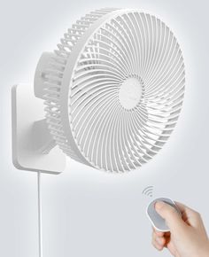 a hand holding a remote control in front of a white wall mounted air vent fan