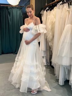 a pregnant woman standing in front of wedding gowns at a bridal shop with her hands on her belly