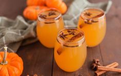 three glasses filled with orange liquid and cinnamon on a wooden table next to small pumpkins