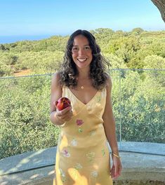 a woman in a yellow dress is holding an apple and smiling at the camera while standing under a tree