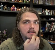 a man with long hair sitting in front of a bookshelf and looking at the camera