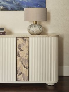 a white cabinet with a painting on the wall behind it and a lamp next to it