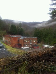 an old building in the middle of a forest with fog rolling over it and cars parked on the road