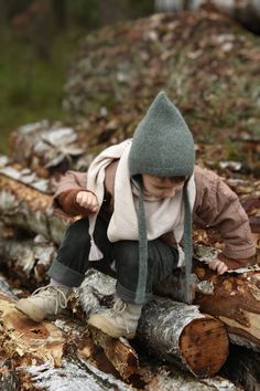 a small child sitting on top of a pile of tree trunks wearing a hat and scarf