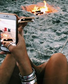 a person sitting in the sand with a cell phone next to a fire pit on their lap